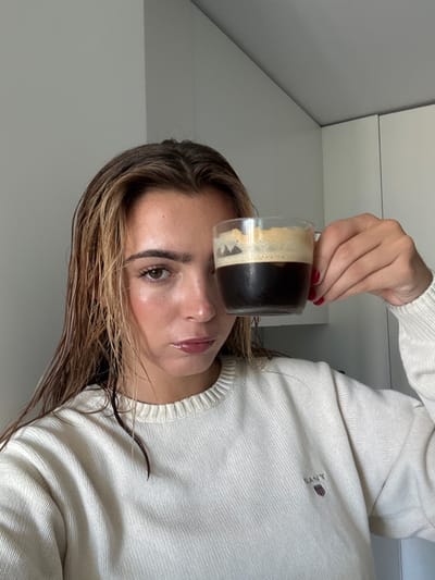 The image captures a female subject from the chest up, positioned indoors. Her light brown hair, appearing damp or wet, is parted centrally. She has visible highlights and wears a cream-colored, ribbed crew-neck sweater with a "GANT" logo, featuring a small shield emblem, on the left chest. Her fingernails are painted red. She holds a clear glass mug containing dark coffee with a thick layer of light brown crema, partially obscuring the right side of her face. Her eyes are dark, and her gaze is directed forward. The background consists of a plain light grey wall on the left and light-colored, possibly white, cabinet doors or wall panels on the right, suggesting an indoor residential or commercial setting. No specific landmarks or unique architectural features are present within the image to pinpoint an exact location within Cidade da Maia, Portugal.