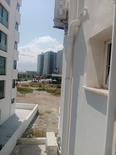 The image shows an outdoor scene in Trikomo, Northern Cyprus, characterized by a mix of ongoing construction and established urban elements under a bright sky with scattered clouds. In the foreground, a section of a modern white building's exterior wall dominates the right side of the frame, featuring a textured finish, a window with a cream-colored frame, and an external white pipe system. To the left, another white building, also with multiple floors and large windows, is partially visible, suggesting a residential or commercial complex.

In the mid-ground, an empty lot with dirt, scattered debris, and patches of dry vegetation is evident. Beyond this lot, a road with several cars, including a white van and other vehicles, indicates traffic flow. The background features a row of tall, modern apartment buildings with numerous windows, appearing to be part of a developed urban area. A construction crane is visible in the far distance, suggesting ongoing development. A large billboard with text and an image, possibly advertising, is also present in the background. The environment appears to be a developing area within a city, with a clear division between newer constructions and undeveloped land.