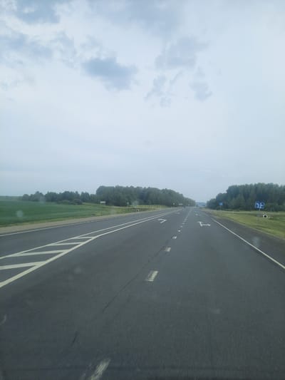 The media file shows a view from a vehicle moving along a paved road. The road is dark asphalt with white lane markings, including diagonal lines marking the right edge of the road and directional arrows painted on the lanes. The road is bordered on the left by a grassy field and a dense line of trees. On the right side of the road, there are two blue road signs with white symbols, one indicating a right turn and the other a straight path with a right turn option. The sky is overcast and light blue with scattered white clouds. The scene suggests a daytime setting, possibly late morning or early afternoon, with moderate weather conditions. The perspective implies a forward-moving viewpoint, characteristic of a dashcam or in-car recording. The image is slightly tilted, giving a dynamic sense of motion. There is no visible text other than the symbols on the road signs. The location context provided indicates Troitskoe, Russia.