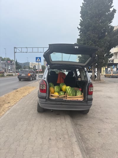 A dark gray car is parked on the side of a road with its trunk open, revealing a large wooden crate filled with watermelons. Several watermelons are also visible outside the crate, some piled on top. An orange garment is draped over a car seat inside the trunk. The scene appears to be on a street with a sidewalk and a road where other cars are visible driving. A pedestrian crossing sign and overhead traffic lights are also present. In the background, there are buildings and trees. The time of day appears to be daytime, and the sky is overcast. The location is identified as Burtaiši, Montenegro.