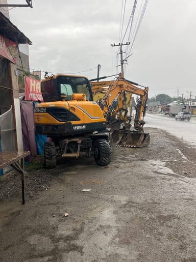 In Medrese, Kyrgyzstan, under an overcast sky, a yellow Hyundai HW60 wheeled excavator is parked on a wet, gravelly roadside. The ground is muddy and appears to have been recently rained upon, with puddles and streaks of water visible. To the left of the excavator, there is a weathered structure with a sloped roof. A red banner with Cyrillic text, partially obscured, hangs from the structure. Also to the left, a simple wooden table with a rough surface stands on the muddy ground. Behind the excavator, another, older Hyundai excavator is partially visible. The road stretches into the distance, lined with utility poles and wires. Several vehicles, including a white pickup truck and a large truck with "PadLease" written on its side, are driving on the wet road. Trees and buildings are visible in the background. The overall scene depicts a construction or work area on a damp, cloudy day.