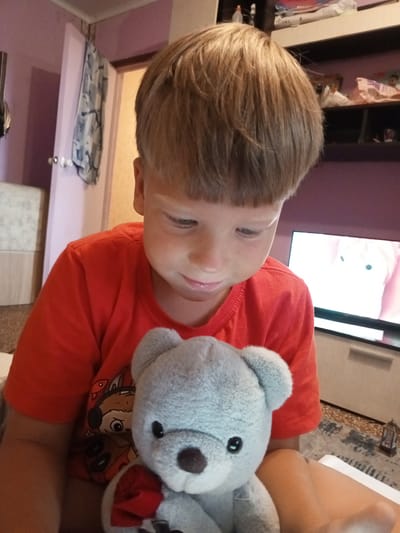 A young boy with short, brown hair and a red t-shirt is holding a gray teddy bear in his lap. The boy is looking down at the teddy bear, and his mouth is slightly open. The teddy bear has black eyes and a brown nose, and it is wearing a red ribbon around its neck. In the background, a television screen is visible, displaying an image of a cartoon character. The room appears to be a living room, with shelves and a dresser in the background. The overall setting is a residential interior, likely a home in the city of Kurchatov, Russia.