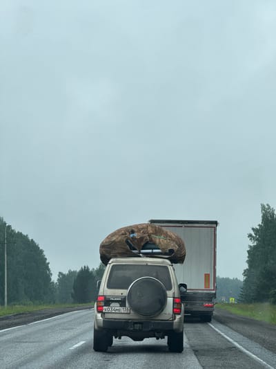 The image depicts a gray, overcast day on a two-lane asphalt road, likely in or near Mar'evka, Russia, given the license plate. The road appears wet, suggesting recent rain. The scene shows traffic, with a white Mitsubishi Pajero in the foreground and a large white truck in the middle distance. The Pajero has a large, tan-colored object covered with a camouflage tarp secured to its roof rack. The license plate of the Pajero is visible and reads "X034ME 138 RUS". The truck in front of the Pajero is a semi-trailer truck, also white, with a red rectangular reflector on its rear. Trees line both sides of the road, and the sky is uniformly gray and cloudy, obscuring any distant features. The overall impression is one of travel on a highway during inclement weather.