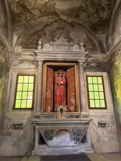 The image shows an interior view of a chapel or alcove, likely within a church, in Ravenna, Italy. The main focal point is a sculptural depiction of Christ lying in repose, dressed in a red robe and adorned with a crown of thorns. The sculpture is encased in a rectangular niche with a gold frame. Above and below the alcove are two windows with multiple panes of glass, emitting green light. The surrounding walls and ceiling are covered in frescoes, adding to the ornate atmosphere. The architectural details of the alcove, including columns and decorative elements, are made of marble. The entire scene conveys a sense of religious reverence and historical significance.