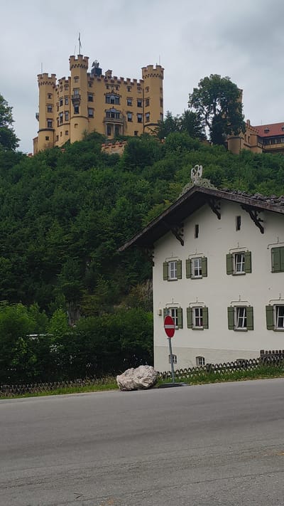 The image shows a yellow castle on a hill covered with dense green trees. In the foreground, a white house with green shutters is visible. A red and white circular sign, likely a "no entry" sign, stands in front of the house. A large white boulder rests on the grassy verge next to a paved road. The sky is overcast. This is likely Hohenschwangau Castle in Schwangau, Germany, known for its picturesque setting and association with King Ludwig II.