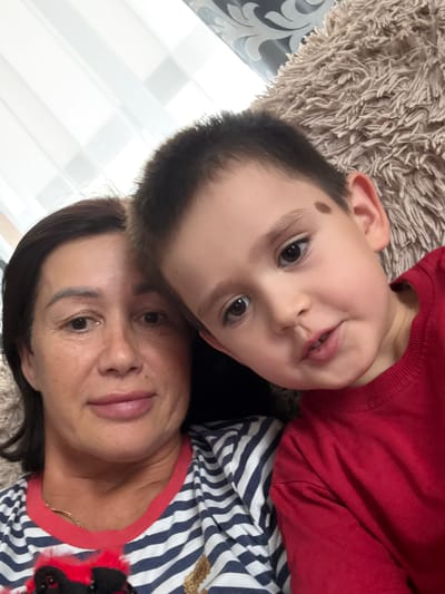 This is a close-up, rotated selfie of a woman and a young boy. They are lying down on a textured, beige rug, with a window and blinds visible in the background. The boy, wearing a red long-sleeved shirt, is positioned above the woman and is looking directly at the camera with a playful expression. He has dark hair and a noticeable brown mole on his forehead. The woman, wearing a striped blue and white shirt with a red collar, is below him and also looking at the camera. Her expression appears relaxed. The lighting suggests it is daytime. The overall scene depicts a candid, affectionate moment between the two individuals.