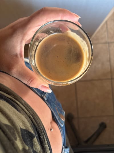 An overhead shot shows a person's left hand holding a clear glass of dark, frothy coffee. The hand is positioned in the upper left quadrant of the frame, with the fingertips and thumb wrapped around the glass. The coffee has a reddish-brown crema on top. The person is wearing a camouflage-patterned shirt and blue denim jeans, with a belly button piercing visible just below the neckline of the shirt. The background consists of a tiled floor with a light brown hue. The scene is indoors, likely in a domestic setting, and there is no indication of the city of Orizari, Bulgaria.