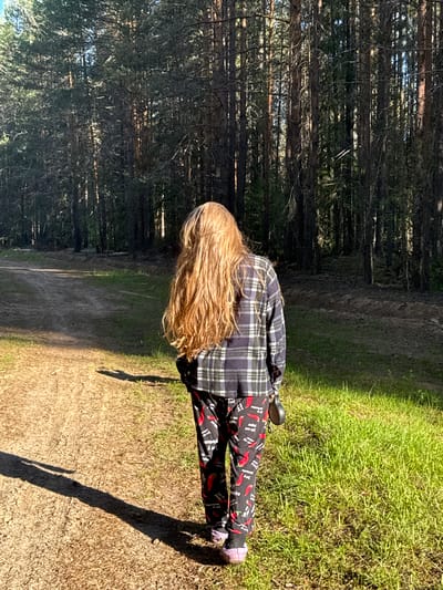 The image, taken in Ust'-Silayka, Russia, shows an individual walking away from the camera on an unpaved dirt path. The person has long, light-colored hair, extending down their back. They are wearing a dark plaid long-sleeved shirt and dark trousers patterned with red chili peppers and white text. Light purple slip-on footwear is visible on their feet. In their right hand, they hold a small, dark, round object. The dirt path shows evidence of use, with visible tracks or impressions. To the left of the path, the ground is primarily bare earth with long shadows indicating strong, low-angle sunlight. To the right of the path, and flanking both sides further back, are areas of vibrant green grass and a dense stand of tall, dark-trunked evergreen trees, consistent with a coniferous forest. The scene is brightly illuminated, with sunlight filtering through the trees.