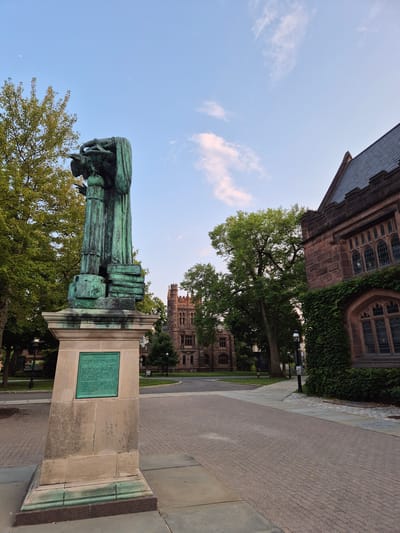 An outdoor, eye-level, wide shot shows a bronze statue in the foreground, a paved walkway, and a historic stone building in the background under a pale blue sky with wispy clouds. The statue, which appears to be an allegorical representation of learning, depicts two figures draped in robes and is mounted on a stone pedestal with a green plaque. The paved walkway is constructed with red-brown bricks laid in a herringbone pattern. To the right, a large, ivy-covered stone building with arched windows and a dark slate roof is visible. In the background, a multi-story stone tower with Gothic architectural elements stands amidst mature trees. The image was taken in Princeton, New Jersey.