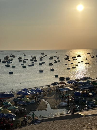 This image depicts a coastal fishing village market scene in Thành phố Phan Thiết, Vietnam, during late afternoon or sunset. The sky displays a warm, golden hue with a bright, low-hanging sun visible in the upper right, casting a shimmering light path across the water.
The sea is populated by over fifty small fishing vessels, including traditional Vietnamese basket boats (thúng chai) and larger motorized fishing boats, scattered across the calm surface. Many of these boats are silhouetted against the bright water.
The sandy beach along the shoreline is densely occupied by a bustling open-air market. Numerous makeshift stalls, covered by a variety of umbrellas predominantly blue, with some green and red, are set up across the sand. People are actively engaged in commerce, moving between stalls, examining goods, and handling items such as white styrofoam containers, plastic tubs, and bundled products. A small channel of water flows across the beach sand.
In the immediate foreground, a concrete or paved embankment slopes downwards from right to left. A single motorcycle is parked on this structure on the left side, and additional individuals are present on the embankment, observing or participating in the market below.