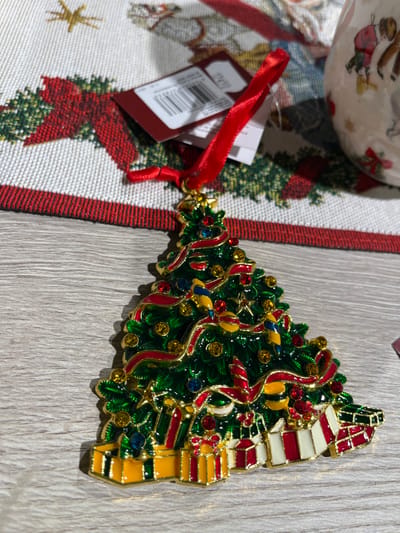 A Christmas tree-shaped ornament sits on a gray wood surface with a holiday-themed tablecloth to the side. The ornament is intricately decorated with gold trim, green enamel, and colorful rhinestones, resembling a Christmas tree adorned with lights, ribbons, and stars. At the top are stacked gift boxes. A red ribbon and tag are attached to the tree’s base. The tablecloth features a white background with a pattern of red poinsettias and green wreaths, accented by a red border.