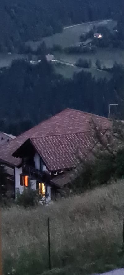 The image shows a house in Deutschnofen - Nova Ponente, Italy, at dusk. The house has a red tile roof and a white facade. There are lights on inside the house, visible through two windows. The house is situated on a grassy slope, with trees and hills in the background. A black metal fence post is visible in the foreground.