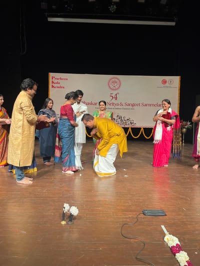 The image shows a group of people on a stage. The stage is made of wood and is decorated with garlands of white flowers. There are two microphones on stands in the center of the stage. The people are dressed in traditional Indian clothing. A man in a yellow and white outfit is bowing to a woman in a red and white sari. There are several other people in the background, including a woman in a blue sari, a woman in a red sari, and a man in a yellow outfit. The stage is decorated with a banner that reads "54th Bhaskar Nritya & Sangeet Sammelan". This event is taking place at Pracheen Kala Kendra in Chandigarh, India (latitude: 30.733, longitude: 76.789).