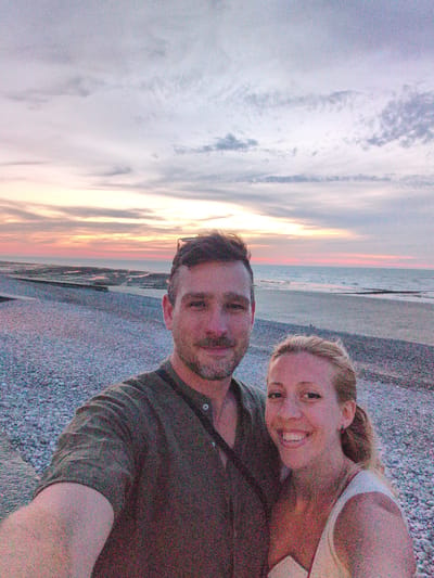 A couple is taking a selfie on a pebble beach at sunset in Veules-les-Roses, France. The man, on the left, has short brown hair and a beard, and is wearing a dark green button-down shirt. The woman, on the right, has blonde hair pulled back and is smiling. She is wearing a white top. The sky behind them is filled with soft pastel clouds, with hues of pink, orange, and purple from the setting sun. The ocean is visible in the background, with gentle waves. The beach is covered in small, smooth pebbles. The overall atmosphere is romantic and peaceful.