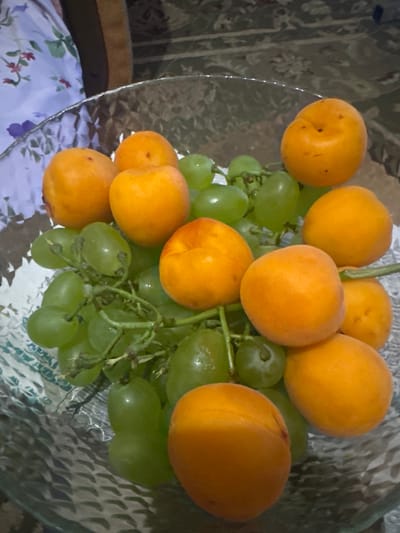 A close-up, overhead shot captures a clear glass bowl filled with ripe green grapes and bright orange apricots. The fruits are arranged artfully, with grapes forming the base and apricots interspersed throughout. The bowl rests on a patterned surface, possibly a tablecloth or a runner, with floral motifs in shades of purple and pink visible in the lower portion of the image. The background is out of focus, showing a textured fabric with a muted, patterned design in neutral tones. The lighting suggests an indoor setting, likely during the day, with a soft, diffused light source. There is no visible text in the image. The overall impression is one of abundance and freshness, highlighting the natural beauty of the fruits. The scene is static, with no discernible activity taking place, implying the image is a still life. The location context of Abay, Kazakhstan, does not offer specific visual cues within the image itself, but provides a geographical backdrop to the setting.