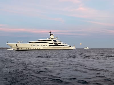 A large, multi-deck, white yacht, equipped with a helicopter on its foredeck, is anchored in a body of water. Other boats, including a sailboat, are visible in the distance. A full moon is visible on the horizon. The sky exhibits hues of pink and blue, suggestive of dawn or dusk. The location is likely the city of Cabo San Lucas, Mexico. The image captures a scene of maritime activity within a serene environment.