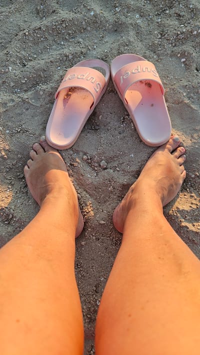 The image depicts a close-up view of two bare feet and a pair of pink slides resting on sandy ground. The sand appears to be mixed with small pebbles and shell fragments. The lighting suggests it is either early morning or late afternoon, casting long shadows. The slides have the word "Super" embossed on them. The scene is set on a beach in Deveses, Spain, indicated by the context provided.