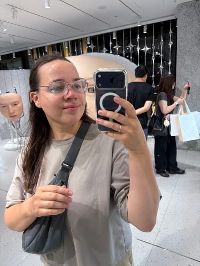 The image captures a woman taking a selfie in what appears to be a well-lit indoor exhibition or retail space, possibly in Chuo, Japan. She is positioned in the foreground, wearing a light-colored t-shirt and a crossbody bag. Her face is framed by silver-rimmed glasses, and she is holding a smartphone to capture the photo. In the background, several art installations or displays are visible, including a manikin head on a stand to the left and a large, abstract sculpture or display behind her. Further in the background, two other individuals are seen, one man in a black t-shirt and a woman holding shopping bags. The environment features clean, modern architecture with white ceilings, speckled floors, and metallic decorative elements on a dark wall. The lighting is bright and even, suggesting an indoor commercial or public space.