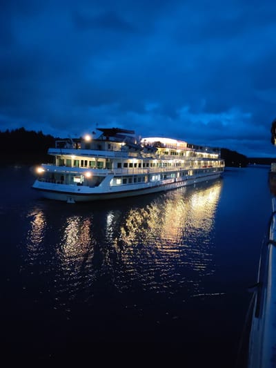 An illuminated river cruise ship travels down a dark river at dusk. The ship is white with multiple decks, each lined with windows. The ship's lights are on, casting a warm glow onto the water. The sky is a deep blue, with some clouds visible. The riverbanks are lined with dark trees. The reflection of the ship's lights shimmers on the water's surface. This scene is taking place in the city of Devyatiny, Russia.