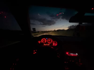 The image captures a view from inside a car at dusk, looking forward through the windshield. The dashboard instruments are illuminated with red light, indicating the car is likely in operation. A portion of the sky with dramatic clouds and a hint of sunset colors is visible through the windshield. The road ahead is visible but obscured by darkness. There are no people visible in the frame. The image appears to be taken in a rural or semi-rural area outside of Aleksandrovo, Bulgaria.
