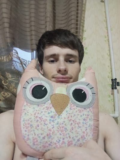 In this close-up, vertically oriented image, a person is holding a plush owl toy. The toy is pink with a floral pattern on its face and has large, round eyes. The person's face is partially visible, and they appear to be looking towards the camera. Their skin tone is fair, and they have dark hair. The background is blurred, showing what appears to be curtains and wallpaper. The overall impression is a casual, indoor setting. There is no visible text in the image. The lighting suggests it could be daytime, but the indoor setting makes it difficult to determine the exact time of day or weather conditions. The person's expression is neutral. The setting seems to be a personal space, likely a home.