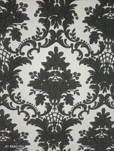 The image displays a close-up, full-frame view of a textured wallpaper with a black damask pattern against a white background. The pattern features ornate, scrolling floral motifs and foliage, with a slightly raised, embossed texture visible on the black elements. There is no discernible location in the city of Улукаўскі сельскі Савет, Belarus, as the image focuses solely on the wallpaper pattern. The composition is static and does not depict any people, actions, or interactions. The lighting is even, highlighting the texture of the wallpaper. A small, partially visible text overlay at the bottom left corner reads "A1 Alpha 20+".