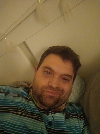 A close-up, indoor photograph depicts a man with brown hair and a beard, wearing a striped blue and black shirt. He is smiling slightly at the camera. The background suggests a bedroom setting with a white wall and what appears to be string lights. The location in Ginsheim-Gustavsburg, Germany is unknown. There is no objective information in the image to infer location, therefore location is an assumption.