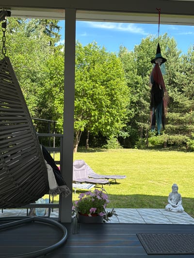 This is an outdoor shot taken from a covered porch, looking out onto a backyard. The porch has dark grey decking and a grey wicker hanging chair with a black and white throw blanket. A potted plant with pink flowers sits on the tiled section of the porch, next to a seated Buddha statue. Beyond the porch, a well-maintained green lawn stretches out, dotted with two lounge chairs. The background is filled with lush green trees, including a conifer on the right. A hanging decoration resembling a witch, with a pointed hat and draped fabric, is suspended from the porch ceiling. The sky is blue with some white clouds. The overall impression is a peaceful, residential backyard in a wooded area. The location of Алешкино Дп, Russia is provided, suggesting a rural or suburban setting.