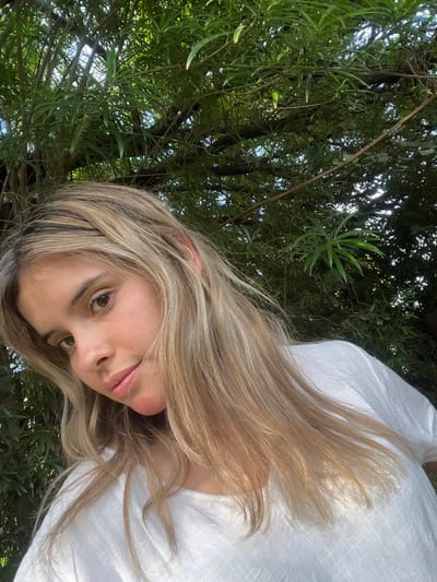 A young woman with long blonde hair and fair skin gazes into the camera with a slight head tilt. She wears a white linen top. Lush green foliage, potentially from a tree, forms the backdrop. The angle is unconventional, suggesting a selfie taken from a slightly lowered perspective, capturing both the subject and the overhead greenery. Sunlight filters through the leaves, creating dappled lighting.
