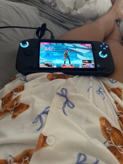 A person is sitting in bed while playing a video game on a handheld console. The person is wearing white pajamas patterned with teddy bears and blue bows. The console is black and has a screen displaying a game with characters and user interface elements. A black cable is plugged into the console. The background shows a bed with gray and white sheets. There is no indication of the city of Casa Blanca, Mexico in the image.