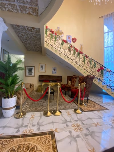 The image shows the interior of a home in Buea, Cameroon. The scene is shot from a low angle, looking up towards a grand staircase decorated with greenery and red ornaments. The staircase is made of white marble and has a wrought iron railing. The room is decorated with a patterned rug and a large potted plant in a white planter. The focal point of the image is a set of four gold stanchions with red velvet ropes. The stanchions are positioned in a semi-circle, creating a barrier across the space. In the background, there are two red velvet armchairs and a table with a flat-screen TV. The home has a modern design with high ceilings and white walls.