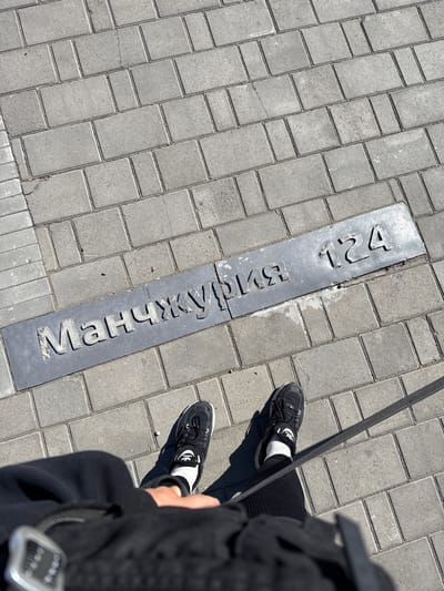 The image shows a section of pavement in Borzya, Russia, with a metal street sign embedded in the brickwork. The sign reads "Манжурия 124" in Cyrillic script, which translates to "Manchuria 124". The pavement consists of grey brick pavers laid in a regular pattern. A person wearing black sneakers with white detailing and white socks is partially visible on the right side of the frame. A black strap or cord extends diagonally from the top right corner of the image, possibly part of a bag or accessory. The environment appears to be an urban area during daylight hours.