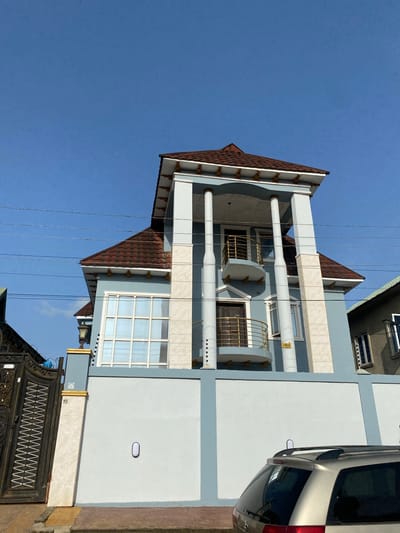 The image depicts a three-story residential building in Idimu, Nigeria. The building features a brown tiled roof, light blue and white exterior walls, and prominent white cylindrical columns. Wrought-iron balconies adorn the second floor. A metallic silver vehicle is partially visible on the right side of the frame. A black metal gate stands in front of the building. Overhead power lines are visible against a clear blue sky. The scene appears to be outdoors during daylight hours.