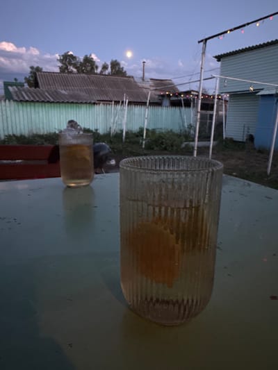 The image depicts an outdoor scene at dusk, possibly in the village of Rodino, Russia. In the foreground, two glasses filled with a light-colored liquid and ice are placed on a green, possibly metallic, table. One glass is in focus, revealing a slice of orange inside. The background shows a rustic setting with a weathered fence, some buildings with corrugated roofs, and trees. The sky is a twilight blue with scattered clouds, and a faint full moon is visible. Decorative string lights are hung above some of the structures, suggesting a festive or social gathering. The overall atmosphere is serene and relaxed, hinting at an evening spent outdoors.