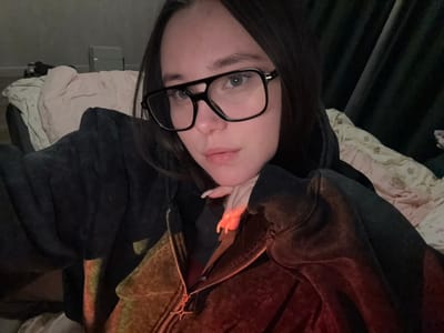 A young adult female is depicted in a close-up, dimly lit indoor setting. She has dark hair and is wearing large, black-framed rectangular glasses. Her expression is neutral, and she looks directly towards the camera. She is wearing a dark, possibly velour or fleece, hooded zipper jacket. A metallic four-pointed star adorns the zipper pull. Her right hand, with manicured nails possibly covered in a light-colored polish, is visible resting beneath her chin, catching a prominent orange-red light reflection that also illuminates parts of her jacket. The background on the left reveals a light-colored patterned comforter on what appears to be a bed or sofa, with a light-toned box object on a surface further back, against a darker wall or curtain. To the right, a dark green or dark grey curtain or fabric is visible. The scene is located in Prikubanskii, Russia.