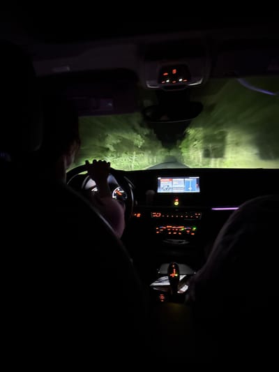 The media file is a low-light, interior shot taken from inside a car at night, rotated 90 degrees clockwise. The scene depicts a driver's perspective.

**Subjects:**
The primary subject is the interior of a car at night, with an emphasis on the dashboard, infotainment system, and steering wheel. A person's hands are visible on the steering wheel, indicating they are driving. Another person's shoulder/head is faintly visible in the bottom left, suggesting a passenger. Outside the car, green foliage is illuminated by the headlights, suggesting trees or bushes alongside the road.

**Activity or event taking place:**
The image captures the act of driving at night. The illuminated dashboard and the headlights casting light on the outside environment confirm the vehicle is in motion.

**Notable details:**
The time of day is clearly night, evidenced by the extreme darkness outside the headlight beam and the illuminated controls within the car. The car's interior is lit by the glow of the dashboard, instrument panel, and infotainment screen. The infotainment screen displays what appears to be a navigation or media interface with a visible "star" icon. On the climate control panel, several orange and red illuminated buttons are visible, along with a digital display showing numbers, possibly a temperature setting (e.g., "22.0"). The green foliage outside suggests the car is traveling through a natural or rural area. Given the location context, this scene occurs in Dulevo, Bulgaria.

**Visible text:**
The infotainment screen shows icons and possibly some unreadable text due to resolution. A "star" icon is prominent. The climate control panel has numbers like "22.0" visible on its digital display. Other illuminated buttons display symbols rather than discernible text.