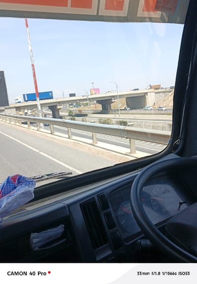 Inside a vehicle, likely a truck, a view through the windshield shows a highway overpass with vehicles on it. A blue truck moves across the overpass. A red and white pole is positioned vertically on the left side of the frame. The dashboard and steering wheel of the truck are visible. The speedometer shows that the truck is running at 70 mph. A piece of cloth rests on the dashboard. The scene is well-lit, suggesting a sunny day.