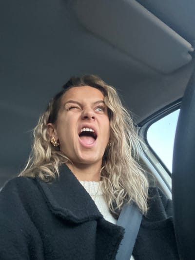 A woman is angled in a car, wearing a seatbelt and a black jacket over a white sweater. She has curly blonde hair and is wearing gold earrings. Her mouth is open in a yell, and one eye is nearly closed, suggesting a grimace or a playful expression. The car interior includes a sun visor and a view of the sky through the windshield.