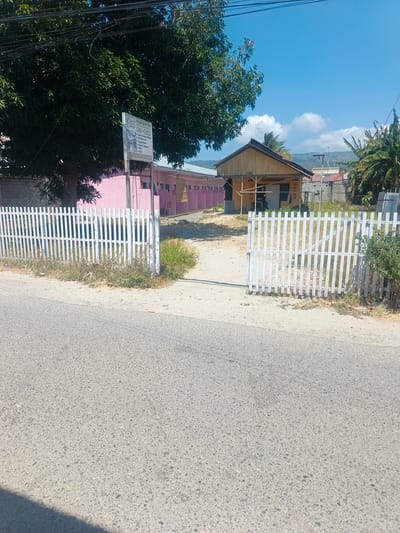 In the city of Tenggela, Indonesia, a street-level view captures a section of a property bordered by a white picket fence. The fence, partially open, reveals a sandy pathway leading towards a building complex. The main building appears to be a single-story structure with a light-colored wooden facade and a brown roof. Adjacent to it, a long, pink building with a darker roof suggests it may house multiple units. A prominent green tree with dense foliage stands to the left of the scene, casting shadows. A white sign with text is affixed to a post near the tree. The environment suggests a warm, sunny day with a clear blue sky and distant hills. The foreground is dominated by a paved road, its surface textured with small stones and gravel.