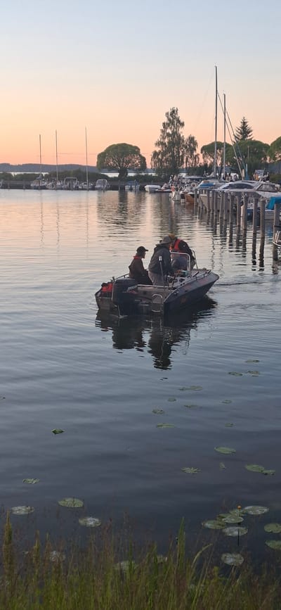 The image depicts a body of water, likely a lake or bay, with several boats docked along a pier on the right side of the frame. The water is calm, reflecting the soft hues of a sunset sky, which transitions from pale pink near the horizon to a light blue at the top. In the foreground, lush green grass and some water lilies are visible along the water's edge.
A small inflatable boat with an outboard motor is moving away from the pier and is the central focus of the image. Three individuals are visible in the boat. One person, wearing a dark cap and jacket, is seated at the rear, facing forward. Two other individuals are positioned in the center and front of the boat, also facing forward. One of these individuals appears to be steering, and the other is looking forward. The boat has a red life vest visible on the back.
In the background, across the water, a tree-lined shore is visible with various types of trees, including some with rounded canopies and others that are more slender. Several sailboats and smaller motorboats are moored at the pier. The overall scene suggests a peaceful evening at a marina or harbor. The location is identified as Salpakangas, Finland, which is known for its lakeside areas and recreational boating activities.