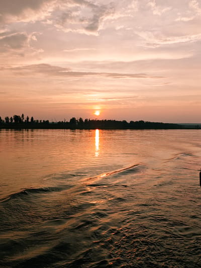 The image depicts a serene sunset over a body of water, possibly a river or lake, in Maloe Nakhval'skoe, Russia. The sky is filled with soft, pastel-colored clouds, predominantly shades of pink and orange, with the sun setting on the horizon. A bright reflection of the sun shimmers on the water's surface. In the distance, a line of trees forms a dark silhouette against the glowing sky, marking the far bank. The foreground of the image shows the rippling surface of the water, with subtle wave patterns suggesting gentle movement. There are no people or man-made structures clearly visible in the scene, creating a tranquil and natural landscape.