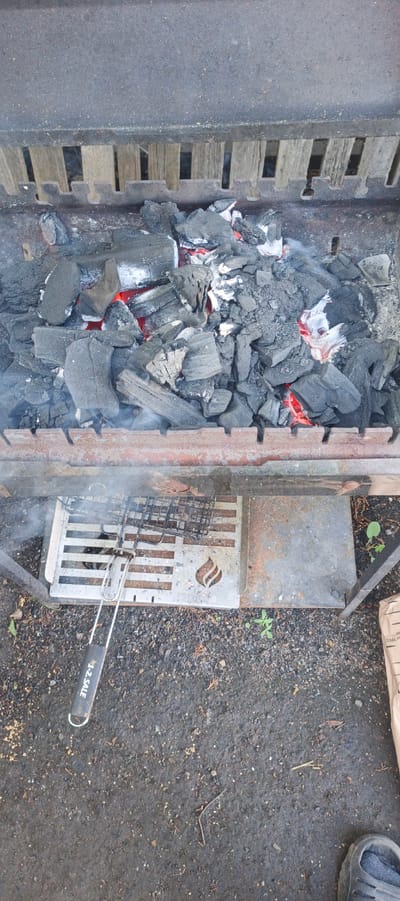 This is an overhead, medium shot of a barbeque grill with glowing charcoal. The grill is filled with chunks of black charcoal, some of which are red hot, indicating that they are burning. There is a light amount of smoke rising from the charcoal. Below the charcoal, there is a metal grate with horizontal bars and a decorative flame cutout. A grill brush with a black handle is placed on top of this grate. The grill itself is made of rusted metal and sits on the ground, which is covered in dirt and small stones. In the background, there are vertical wooden slats, likely part of the grill's construction. To the right of the grill, a small green plant is visible growing in the dirt. The overall scene suggests an outdoor setting where a barbeque is being prepared or has recently been used. The lighting appears to be natural daylight. Visible text on the grill brush reads "1-2. SALE".