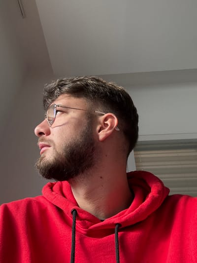 A close-up, profile image depicts a young adult male, positioned indoors in Lièpvre, France, looking towards the left side of the frame. The subject has short, dark brown hair with faded sides and a trimmed beard and mustache. He wears round, thin-framed eyeglasses with clear or lightly tinted lenses and a red hooded sweatshirt with visible black drawstrings at the collar. A strong light source, likely from a window, illuminates the left side of his face, casting a shadow on his right cheek and neck. The background consists of a light-colored ceiling and an upper portion of a wall on the left, and part of a window with horizontal blinds on the right. No other individuals or specific objects are identifiable.
