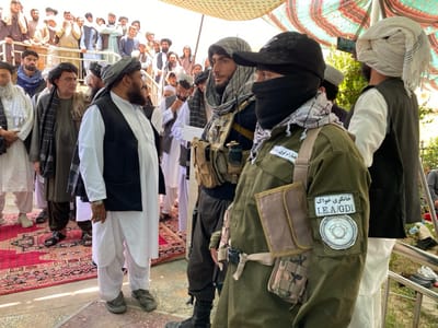 The image depicts a group of men, some wearing traditional Afghan attire, in the city of Zaker-E Sharif, Afghanistan. The men are gathered under a canopy, seemingly in a public space. Two men, wearing military fatigues and black masks, stand towards the right of the image. One of them is adorned with a patch that reads "I.E.A./GDI" in English and "خاطری جرات" in a foreign script. The men are likely armed with firearms, though these are not visible in the image. The men in the background, many wearing white turbans, are engaged in conversation, seemingly unconcerned by the presence of the armed men. The environment suggests a public gathering or event in Zaker-E Sharif.  The image is taken from a low angle, giving the viewer a perspective from the ground. The overall atmosphere is one of normalcy amidst a visible security presence.