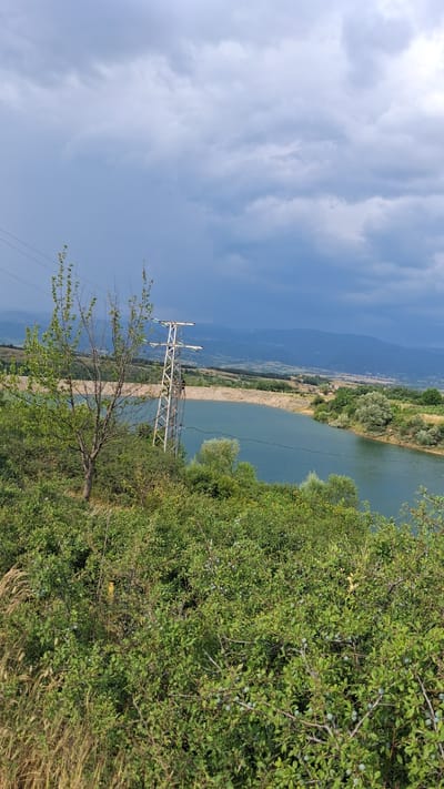 This media file depicts a landscape scene in Dobrotino, Bulgaria. The image is oriented vertically. In the foreground, there is lush green vegetation, including bushes with small green fruits, and a bare tree with a few leaves. A tall, white metal electricity pylon with wires is visible in the middle ground. Beyond the vegetation and pylon, a wide, calm body of water, likely a river or lake, stretches across the scene. The water is a tranquil shade of blue-green.

On the far side of the water, there is a shoreline with more greenery and some trees. In the distance, a valley unfolds with rolling hills and mountains. A small settlement with scattered buildings can be seen nestled in the valley. The sky is partly cloudy, with patches of bright blue interspersed with soft, white clouds. The lighting suggests it is daytime, likely late morning or early afternoon, with clear, fair weather. The overall impression is one of a serene, natural landscape enhanced by human infrastructure. There is no visible text in the image.