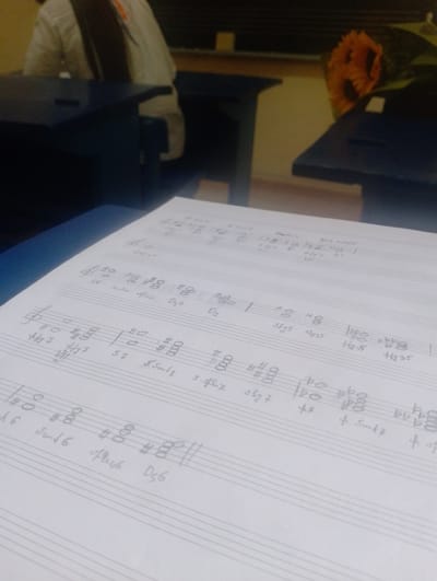 The image shows a close-up view of sheet music with handwritten notations, resting on a blue surface, possibly a desk. A person wearing a white shirt and dark hair is partially visible in the background, seated at a desk. The overall setting appears to be indoors, with a blurred background that includes what might be a bouquet of sunflowers on a desk. The sheet music itself features musical staves, clefs, note heads, and various symbols and text, suggesting a musical composition or study. The lighting is dim, casting a shadow on the sheet music.
