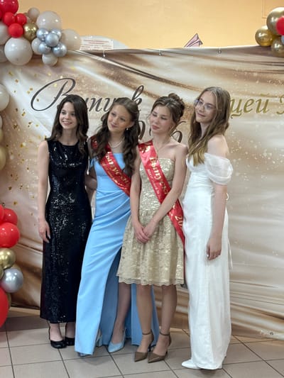 This media file captures a celebratory moment with four young women dressed in elegant gowns, posing together in front of a decorative backdrop. The backdrop features a shimmering, abstract design with large, ornate cursive text that appears to be in Russian. Banners with "Выпуск" (Vypusk - Graduation) and "20" are visible, suggesting the event is a graduation ceremony or related celebration.
The women are posed in a staggered, standing arrangement. From top to bottom, the first woman wears a sparkly white off-the-shoulder gown. The second is in a cream-colored lace dress with a red sash that reads "Выпуск МБОУ лицей № 33" (Graduation MBOU Lyceum No. 33). The third wears a light blue floor-length dress and also has a red sash. The fourth, at the bottom, is in a striking black sequined dress. They all appear to be in good spirits, with smiles and relaxed postures.
To the left of the women, a balloon arch in shades of cream, gold, silver, and red adds to the festive atmosphere. The floor appears to be tiled. The lighting is bright, suggesting an indoor setting, possibly a school hall or event space. The overall impression is one of joy and accomplishment, likely marking the end of a significant academic chapter for these young women. The presence of the specific school name on the sashes indicates a local event.