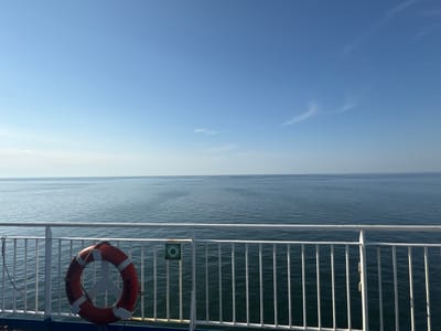 This is a photograph taken from the deck of a ferry, looking out over a calm, blue ocean under a clear sky. The horizon line is sharp and distinct, with the vast expanse of water meeting the pale blue of the sky. A white railing runs along the foreground, with vertical white bars creating a repetitive pattern.

Hanging on the railing to the left is a bright orange life preserver. Text is visible on the life preserver, reading "ROBIN HOOD" and "EMDEN" in white lettering. To the right of the life preserver, a small green circular sign with a white symbol is mounted on the railing.

The lighting suggests it is daytime, possibly morning or late afternoon, with the sun casting a gentle illumination on the scene. The water is largely smooth, with only slight ripples, indicating calm weather. The overall mood of the image is serene and peaceful, evoking a sense of journey and open space. The location context is unknown, with only Lithuania provided as a possible hint.