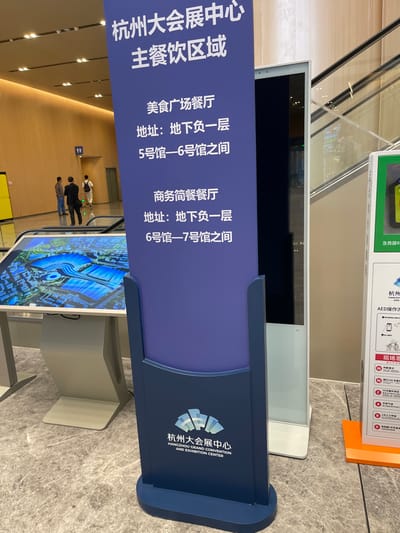 The scene is a brightly lit indoor hallway in the Hangzhou Grand Convention and Exhibition Center. It is located in Hangzhou, China, at 30.273° N, 120.416° E. Two men are walking in the hallway. On the left, a large digital display with a map shows the layout of the convention center. On the right, a large digital display shows a blank screen. A tall, freestanding sign stands in the center of the hallway. The sign is a dark blue color and features the words "Hangzhou Grand Convention and Exhibition Center" in English. The sign also indicates the location of the "Beauty Salon" and "Commercial Bank", both located on the lower floor. To the right of the sign, there is a large, bright orange sign with the words "AED" and "Emergency Medical Services" in English. The signs are positioned in a way that would likely be easily visible to visitors.