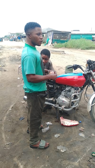 The image depicts a scene on a dirt road, likely in Warri, Nigeria. Two individuals are present, focused on a red and blue motorcycle. The person in the foreground, a young man wearing a teal t-shirt and dark pants, stands looking to the right, his hands near the motorcycle's fuel tank. Another person, whose face is partially visible, is behind the motorcycle, also appearing to be engaged with it. The motorcycle has a chrome engine and a red and blue fuel tank. The environment is characterized by a dusty, uneven ground with scattered debris, including plastic wrappers and a red plastic object. In the background, there are makeshift structures, possibly shops or stalls, and other vehicles, suggesting a commercial or roadside area. The sky is overcast.
